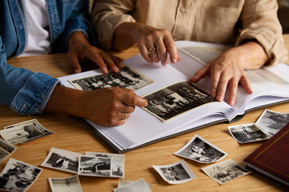 Two people placing old black-and-white photographs into a photo album on a wooden table.