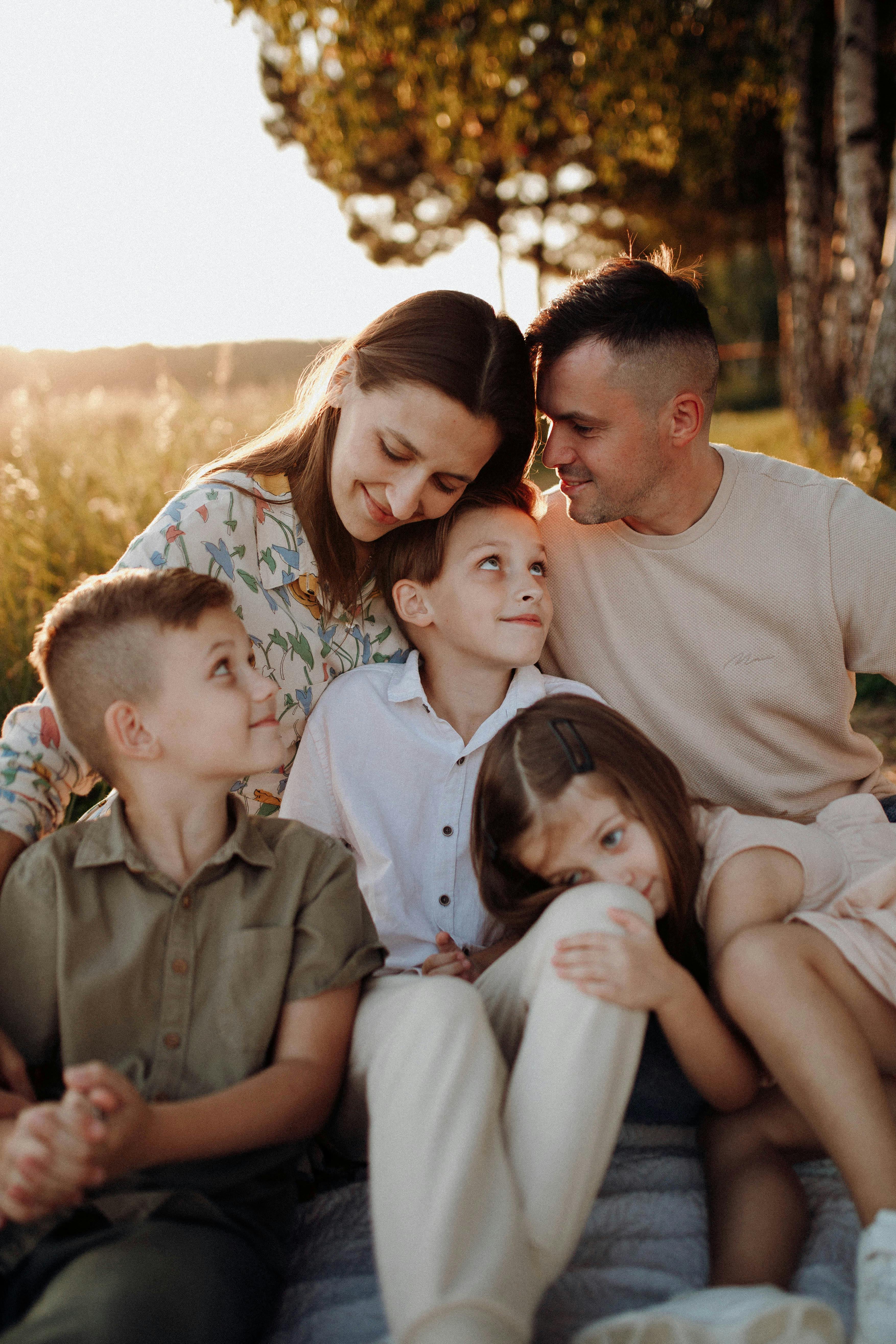 A smiling family of five sitting closely outdoors in warm sunlight, with two parents and three children cuddling together.